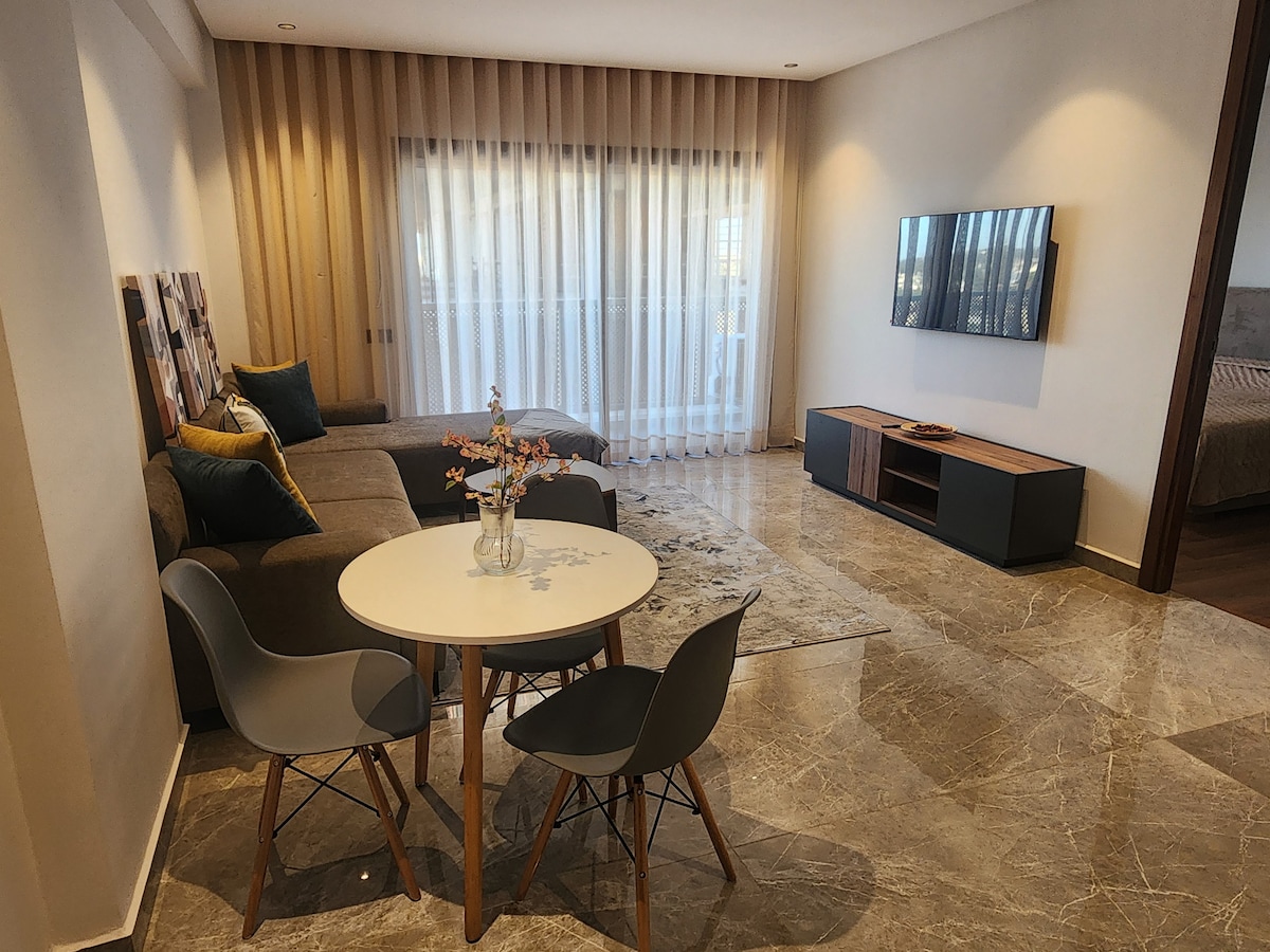 A modern living area features a comfortable sofa alongside a round dining table with four chairs. Light streams through floor-to-ceiling sheer curtains, illuminating the space. A flat-screen TV is mounted on the wall, and the flooring is composed of elegant marble tiles.
