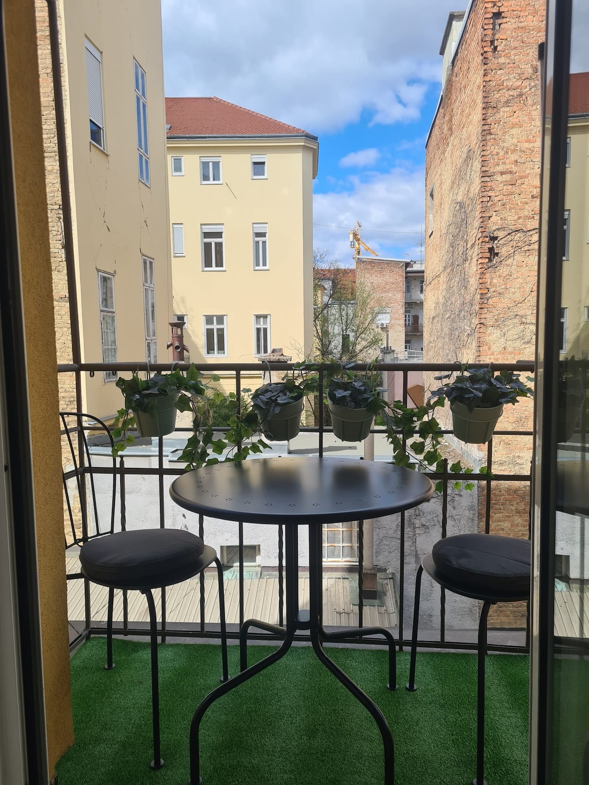 A small balcony is visible, featuring a round black table and two black chairs. Green foliage in planters adds a touch of nature. The view outside includes nearby buildings and a blue sky with fluffy clouds.
