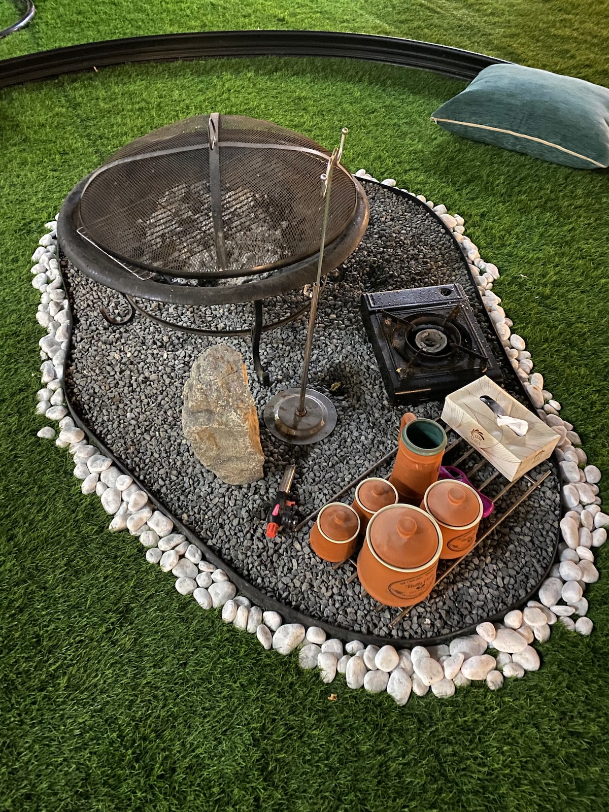 A fire pit area is surrounded by decorative pebbles and artificial grass. The fire pit features a metal grate, while a gas stove stands nearby. Several terracotta pots and a lighter are arranged next to a rock, inviting cozy gatherings in a serene outdoor setting.