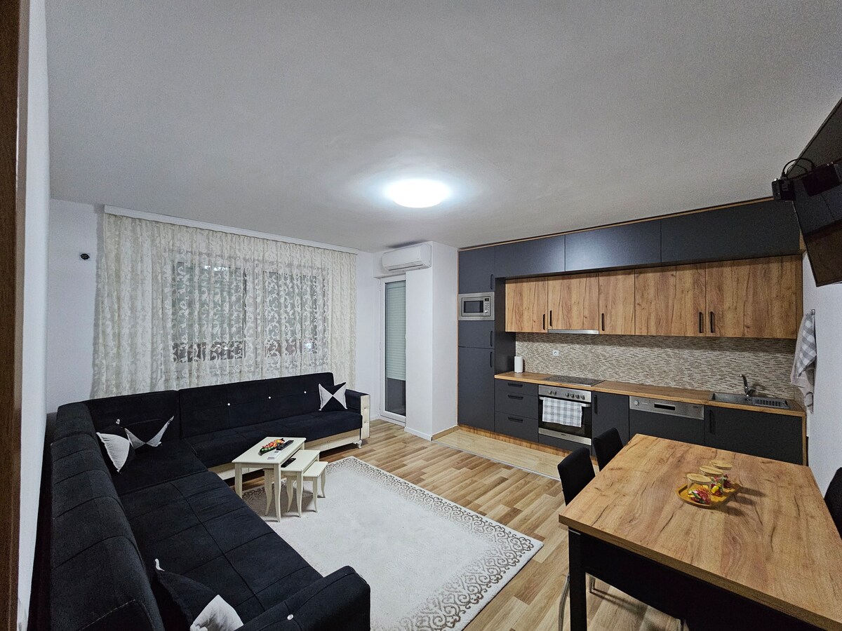 The spacious living area features a large, dark sectional sofa and a light dining table. A well-equipped kitchen is showcased with wooden cabinetry and a modern design. Natural light filters through sheer curtains, illuminating the room's neutral tones and enhancing the overall ambiance.