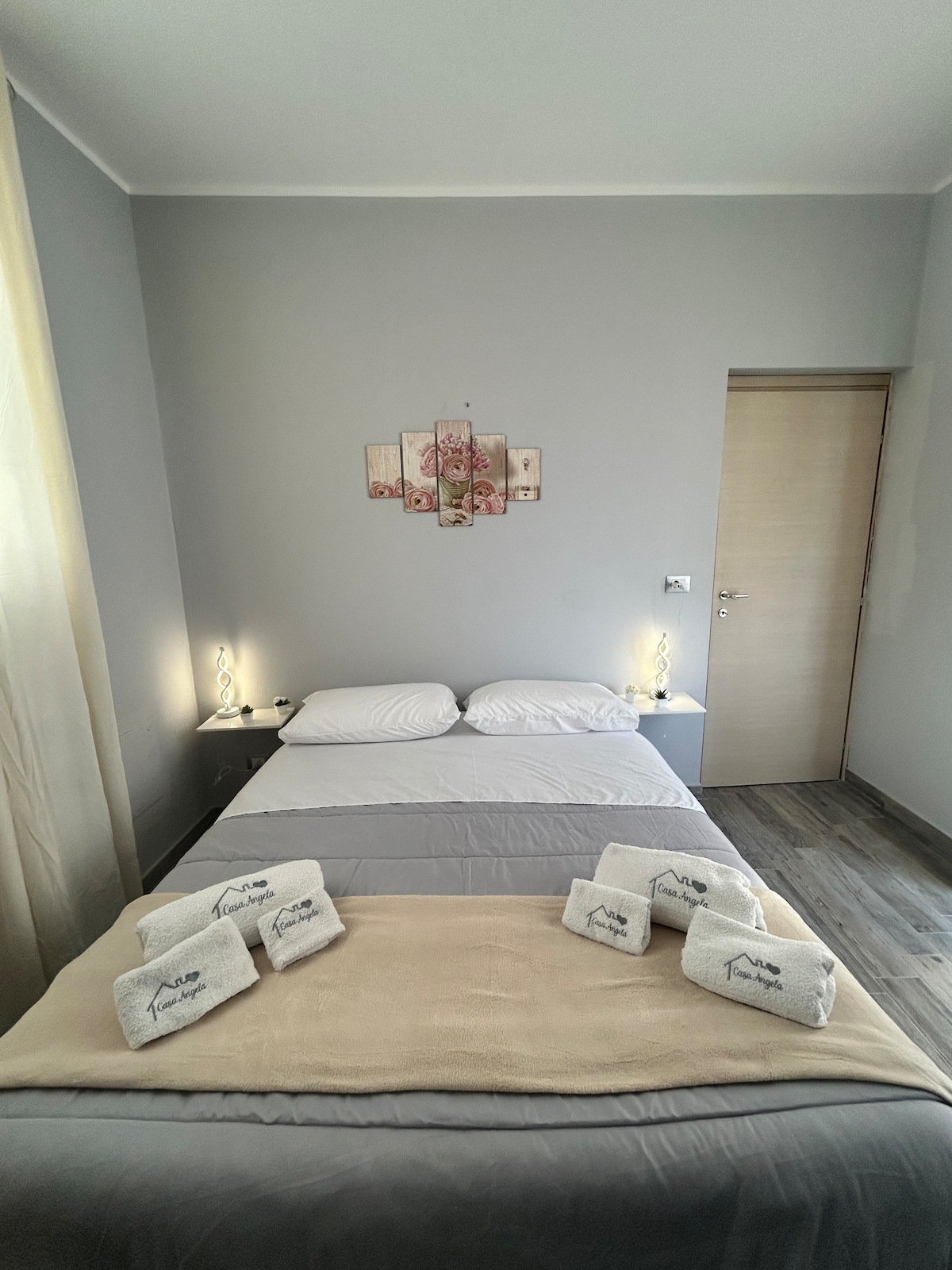 A calming bedroom features a spacious bed with neutral-colored bedding, complemented by soft lighting from bedside lamps. Decorative wall art adds a touch of elegance, while the room's light-colored walls enhance its airy feel. Towels are neatly arranged on the bed, ready for use.
