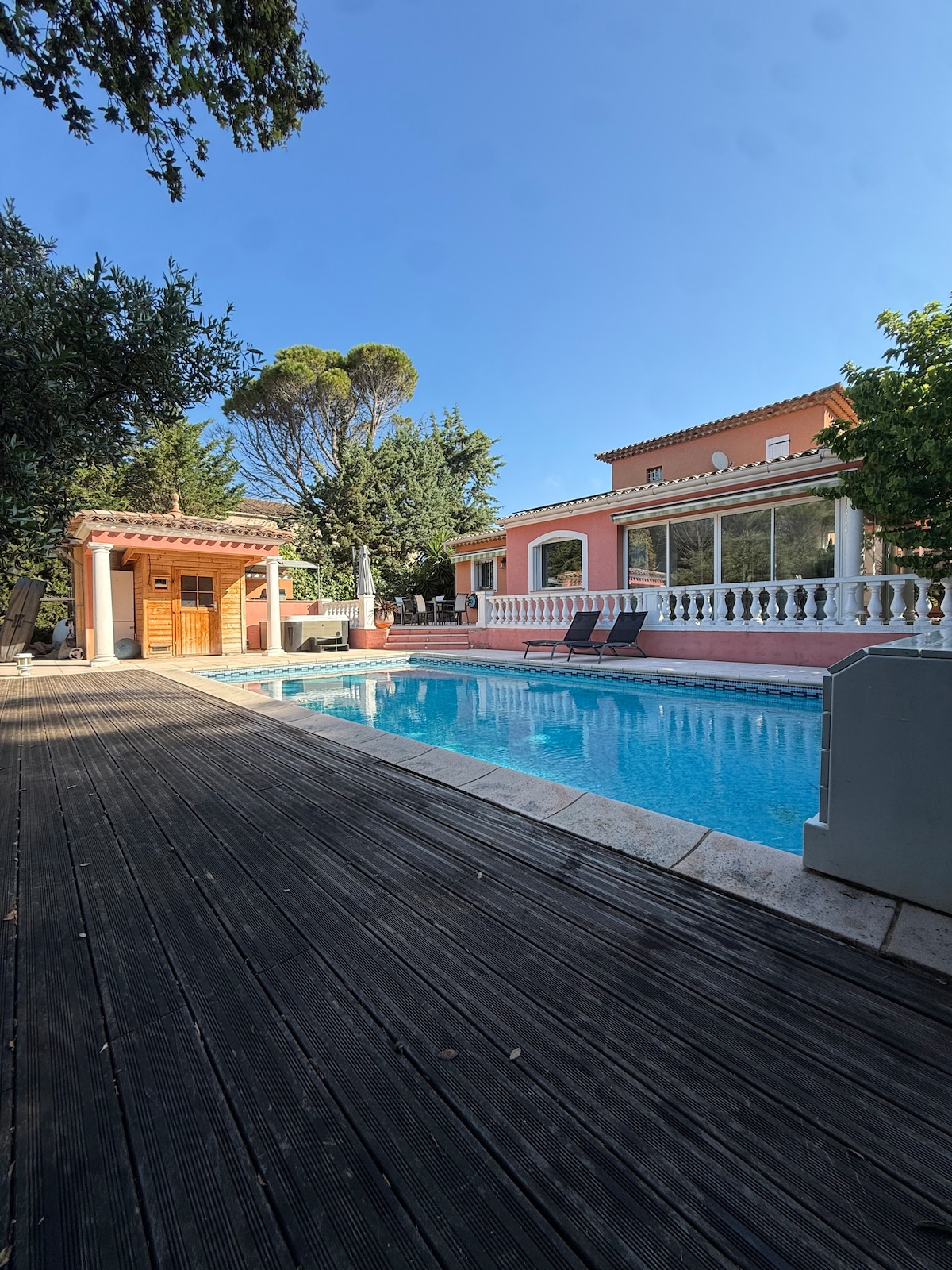A serene outdoor area features a swimming pool surrounded by a wooden deck. Two lounge chairs are positioned near the water, and a nearby wooden structure suggests sauna facilities. Lush greenery and trees provide natural shade, and the villa's exterior highlights soft pink tones.
