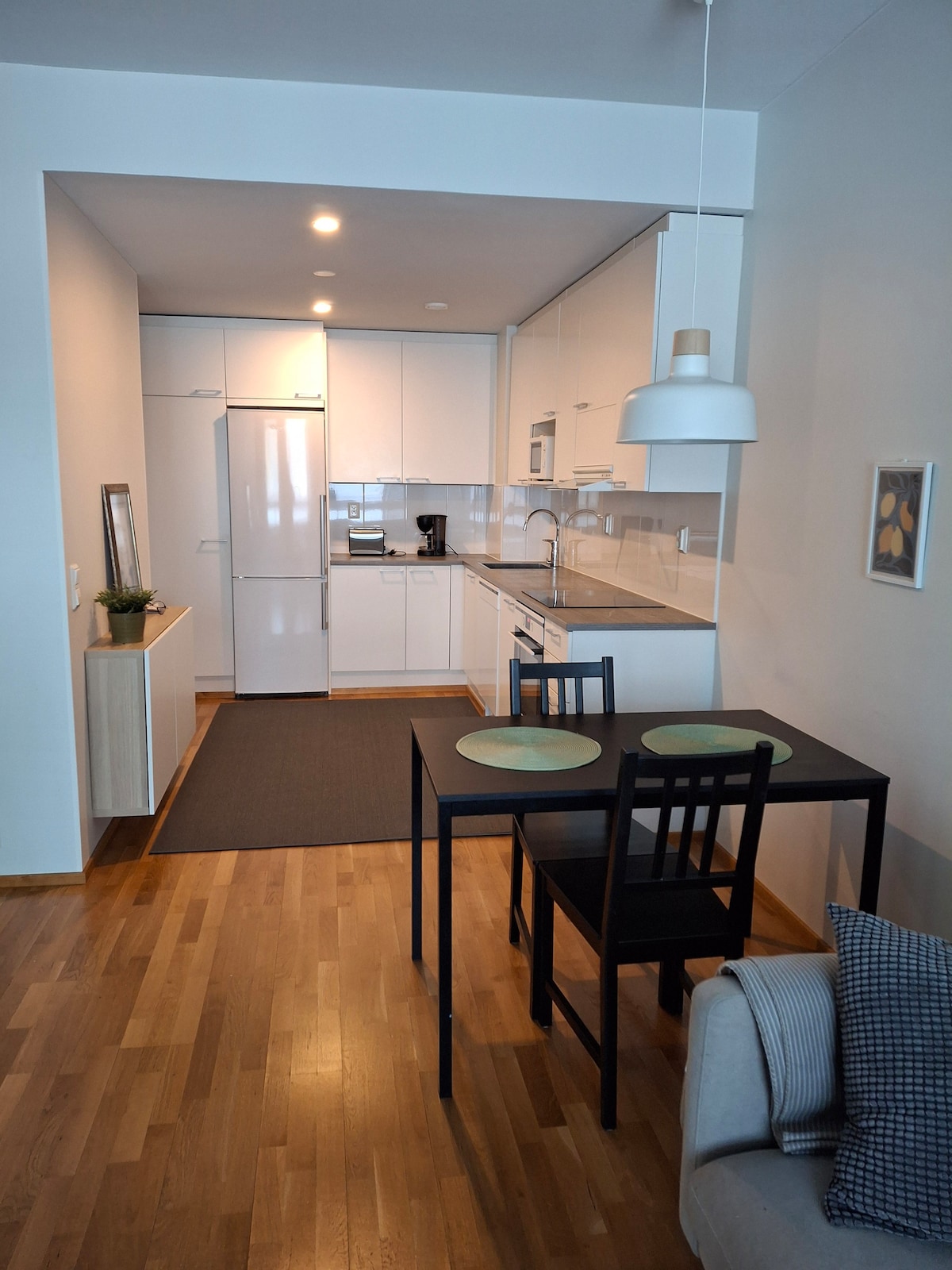 An open-plan kitchen and living area is displayed, featuring modern white cabinetry and appliances. A small dining table with two chairs is positioned in front of the kitchen, while a cozy seating area is situated nearby. Light wood flooring enhances the room's overall brightness.