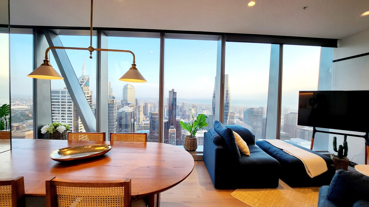 A modern dining area features a round wooden table surrounded by woven chairs, complemented by a pendant light overhead. A cozy seating arrangement with a blue sofa and a white throw is positioned near floor-to-ceiling windows, showcasing expansive views of the Melbourne skyline.