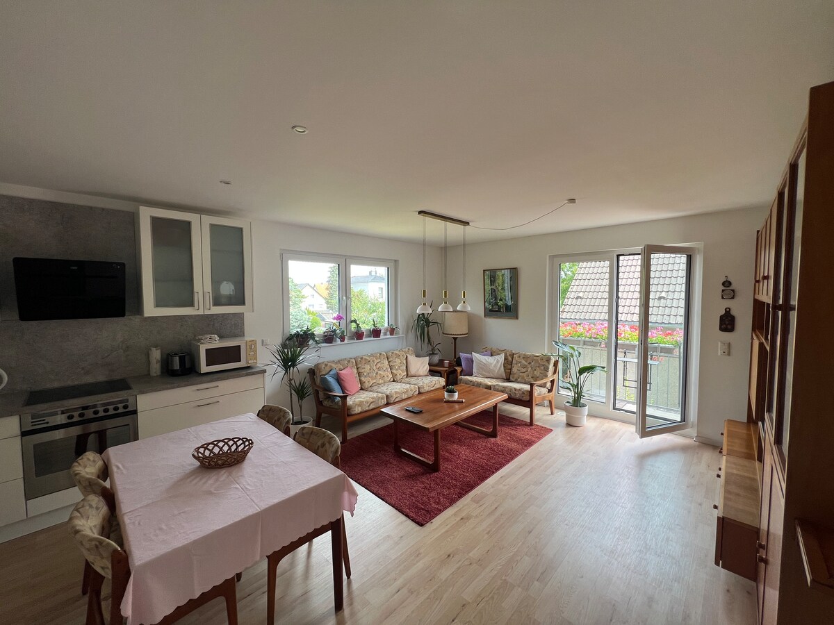 A spacious living area is presented with a dining table set for four, alongside a cozy seating arrangement featuring two sofas. Large windows allow natural light to fill the space, with a view of a balcony adorned with flowers. A modern kitchen area is visible in the background.