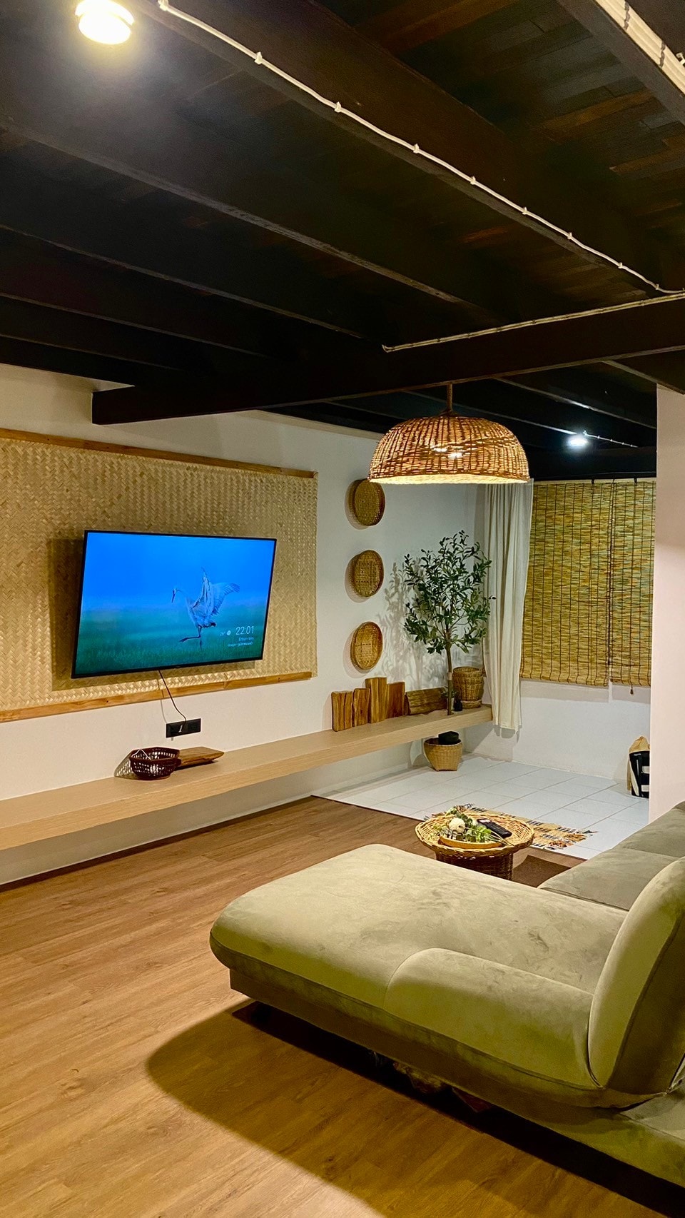 The living room features a spacious sofa facing a wall-mounted 55-inch Smart TV. Natural light enters through a partially covered window, complemented by decorative wall elements and a potted plant. The wooden ceiling beams enhance the rustic charm of the space.
