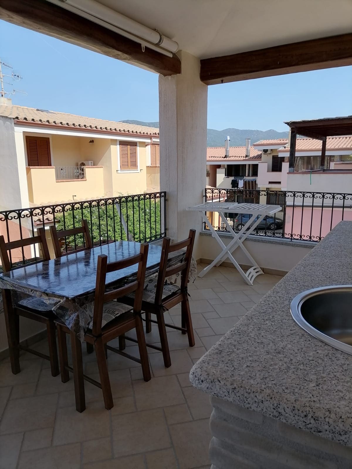 A spacious terrace features a dining table with four chairs and a countertop with a sink. An outdoor clothing drying rack is positioned nearby. A view of neighboring houses and distant mountains is visible, framed by the terrace's railing.