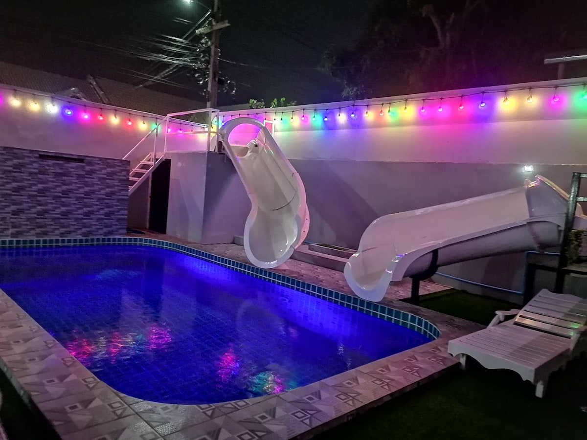 An illuminated swimming pool features two white water slides positioned at one end. Colorful lights line the surrounding walls, creating a vibrant atmosphere at night. Lounge chairs are placed nearby, offering a space for relaxation.