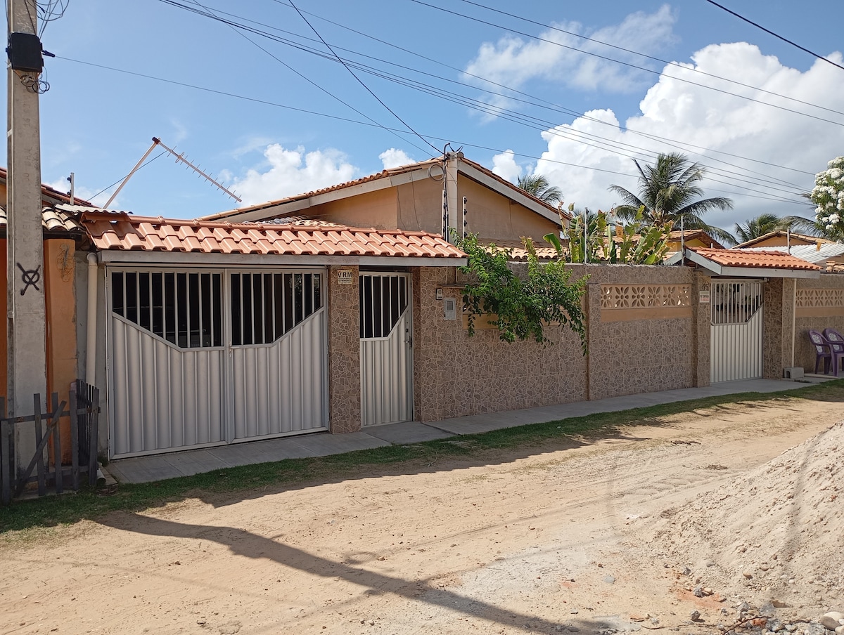 A residential structure features a combination of stone and stucco walls with a terracotta tile roof. Two white metal gates are visible, flanked by low decorative concrete walls. The surroundings include sandy pathways and tropical vegetation under a bright blue sky.