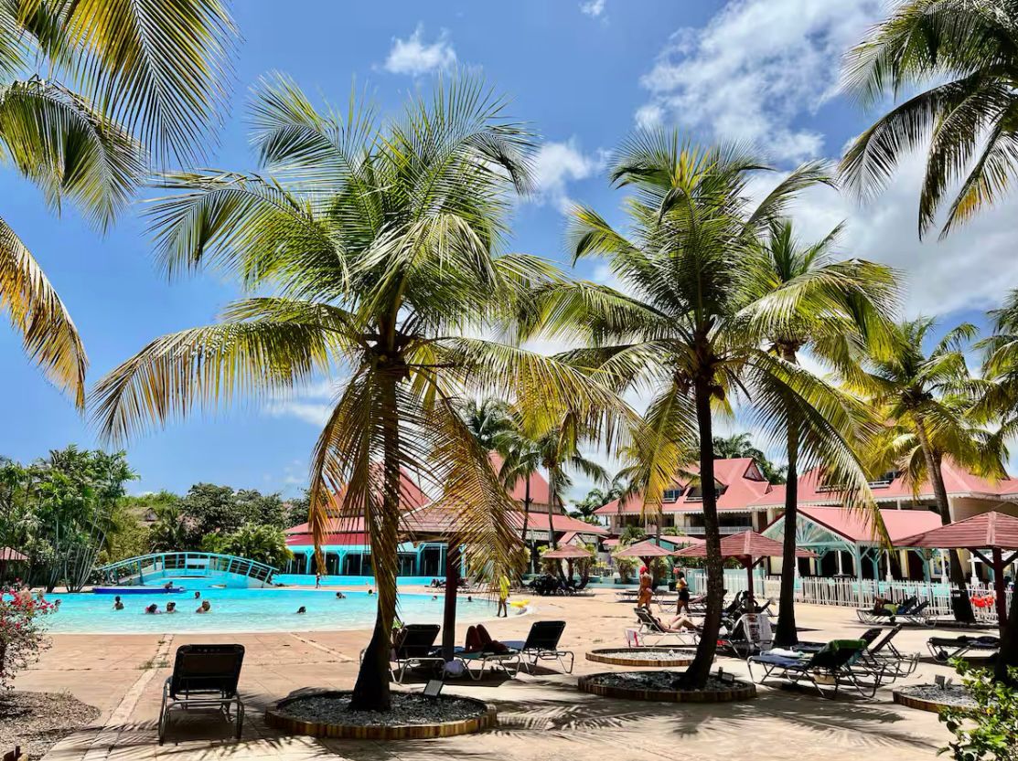 A sandy area is framed by palm trees swaying gently in the breeze, overlooking a large swimming pool. Lounge chairs are arranged near the water’s edge, with colorful umbrellas providing shade. The setting features a lively atmosphere with people enjoying the facilities.