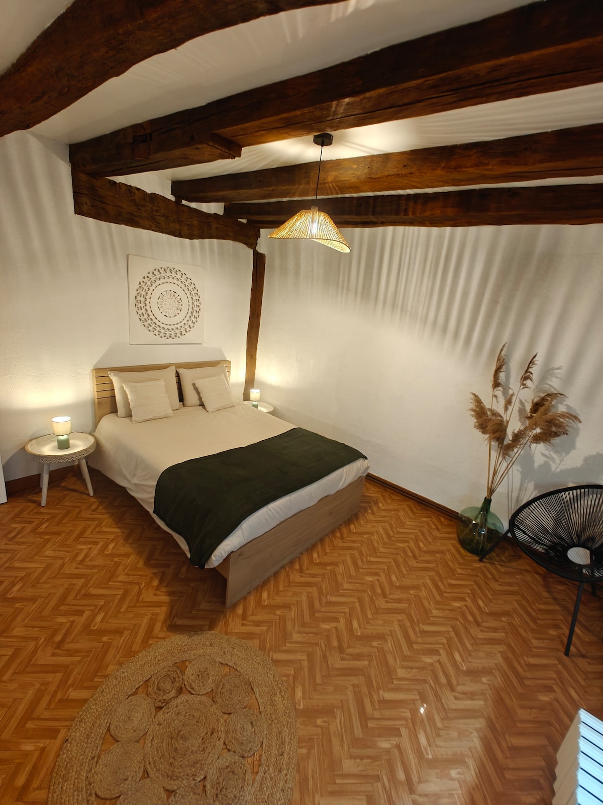 A spacious bedroom features a large bed with neutral linens and decorative pillows. Exposed wooden beams add character to the high ceiling. Soft lighting is provided by a pendant lamp, and natural elements are present with a plant and dried pampas grass in the corner.