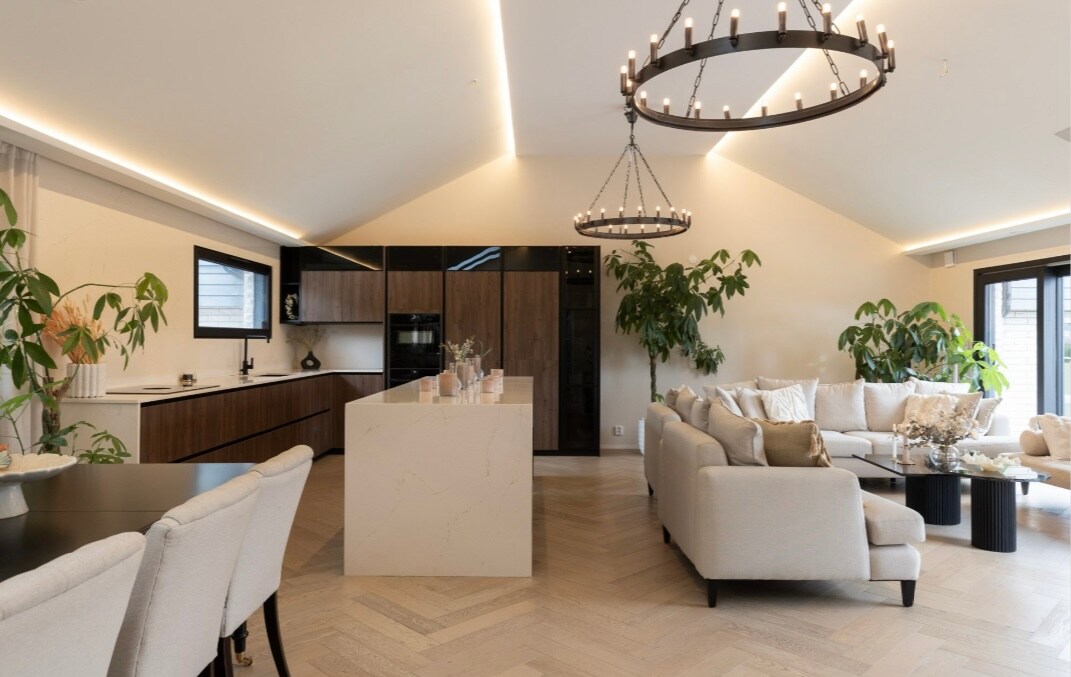 An open-plan living area features high ceilings and large windows, allowing natural light to fill the space. A modern kitchen with dark wood cabinets and a white island complements a well-arranged seating area with a comfortable sofa and decorative plants.