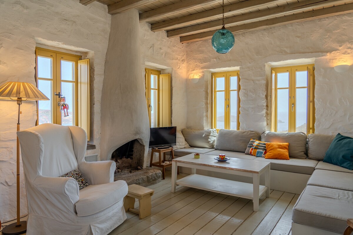 A living room features soft seating arrangements, including a cozy armchair and an expansive sectional sofa. Natural light streams through multiple yellow window frames, highlighting the textured stone walls. A simple coffee table is centered in the space, with a modern light fixture suspended from the ceiling.