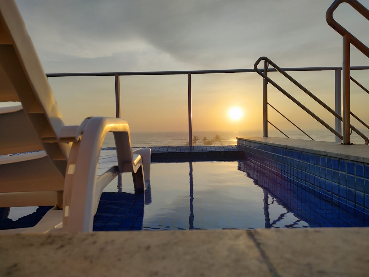 A serene infinity pool reflects the soft hues of a sunset, offering a view of the ocean's horizon. Lounge chairs are positioned nearby, providing a spot to relax and enjoy the tranquil scene.