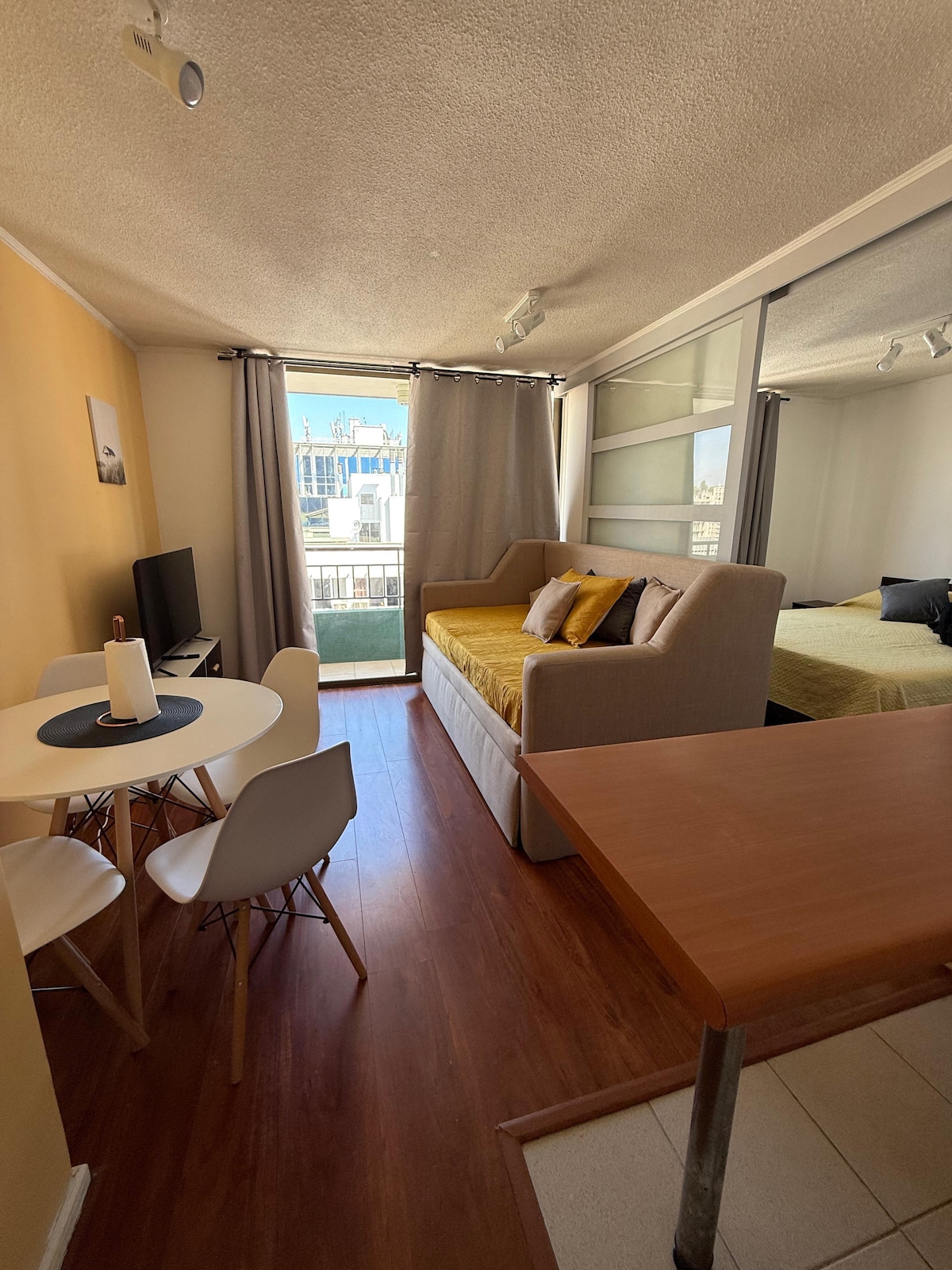 A comfortable living area is showcased, featuring a light-colored sofa positioned against a wall. A dining table with four chairs is visible, alongside a TV unit. Natural light floods the space through large windows, which offer a view into the outdoors, enhancing the inviting settings.