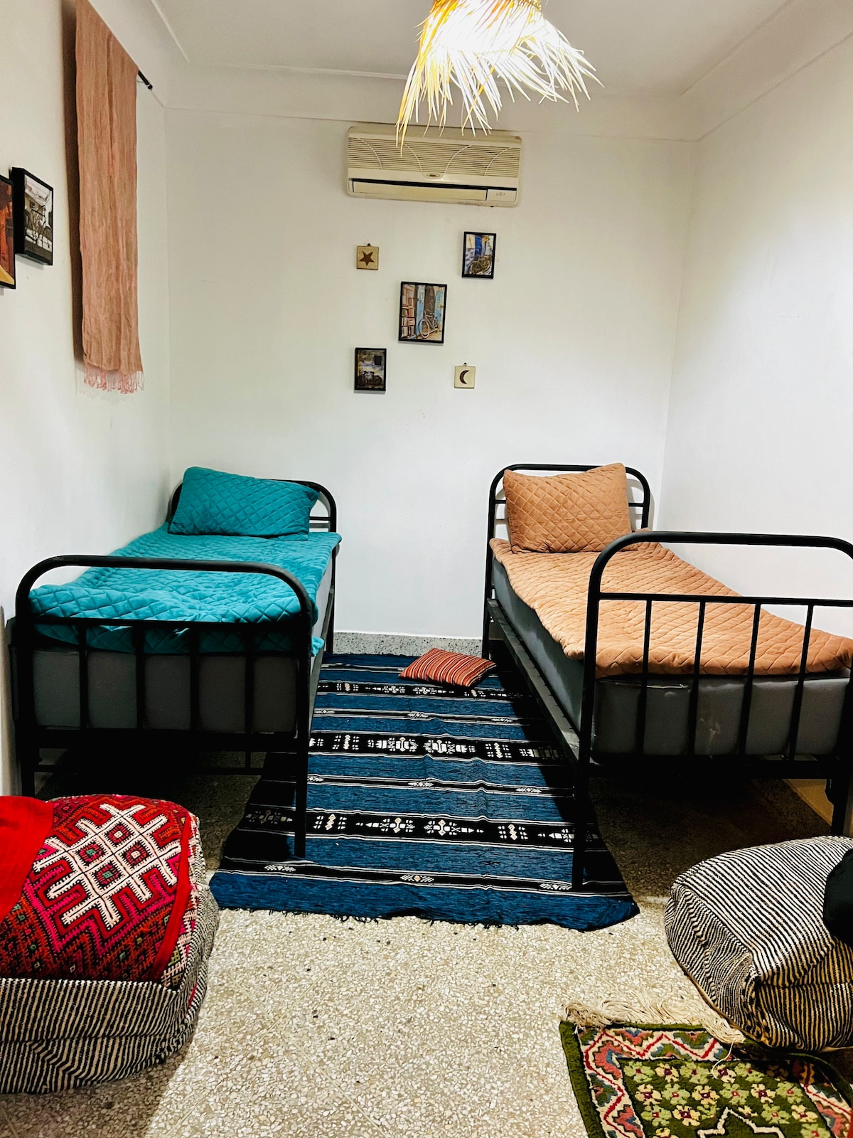 A simple sleeping area is furnished with two metal-frame beds, one covered with a blue quilt and the other with a tan coverlet. A decorative rug lies on the floor, adding texture, while vibrant wall hangings and framed pictures decorate the pale walls.