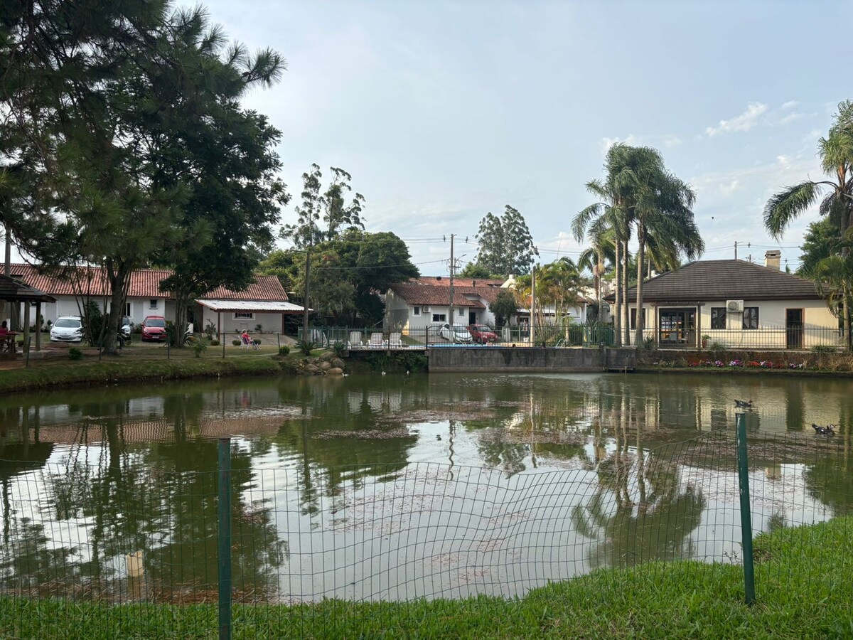A serene pond reflects surrounding greenery and structures, with subtle ripples on the water's surface. Lush grass lines the pond's edge, and various trees provide shade. Nearby, a few modest buildings are visible, creating a peaceful community ambiance.