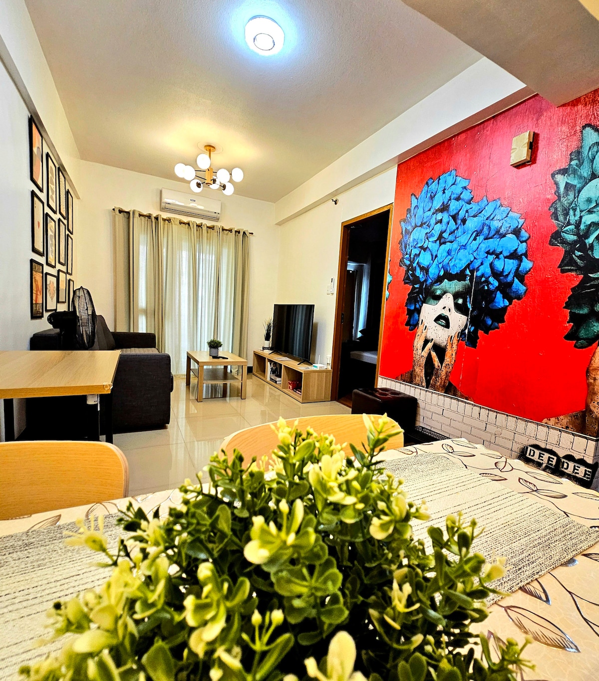 A modern living area is highlighted with a vibrant mural featuring blue trees. A comfortable sofa sits next to a television, while a wooden dining table adorned with a decorative plant is positioned in the foreground. Natural light enters through partially draped windows.