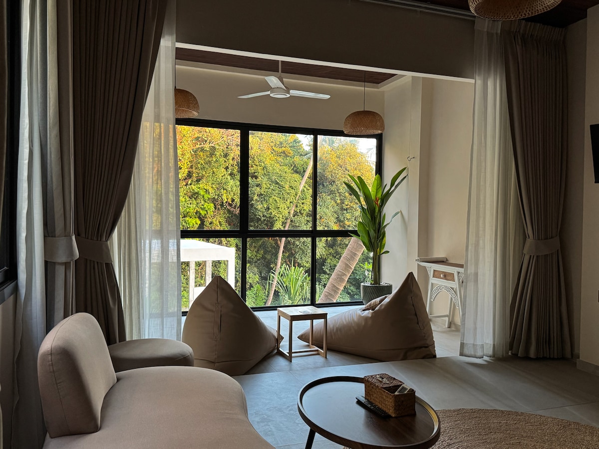 A spacious living area features large windows that overlook a vibrant jungle canopy. Soft beanbag chairs are placed beside a round coffee table. Light-colored curtains frame the windows, while a green potted plant adds a touch of nature to the interior.