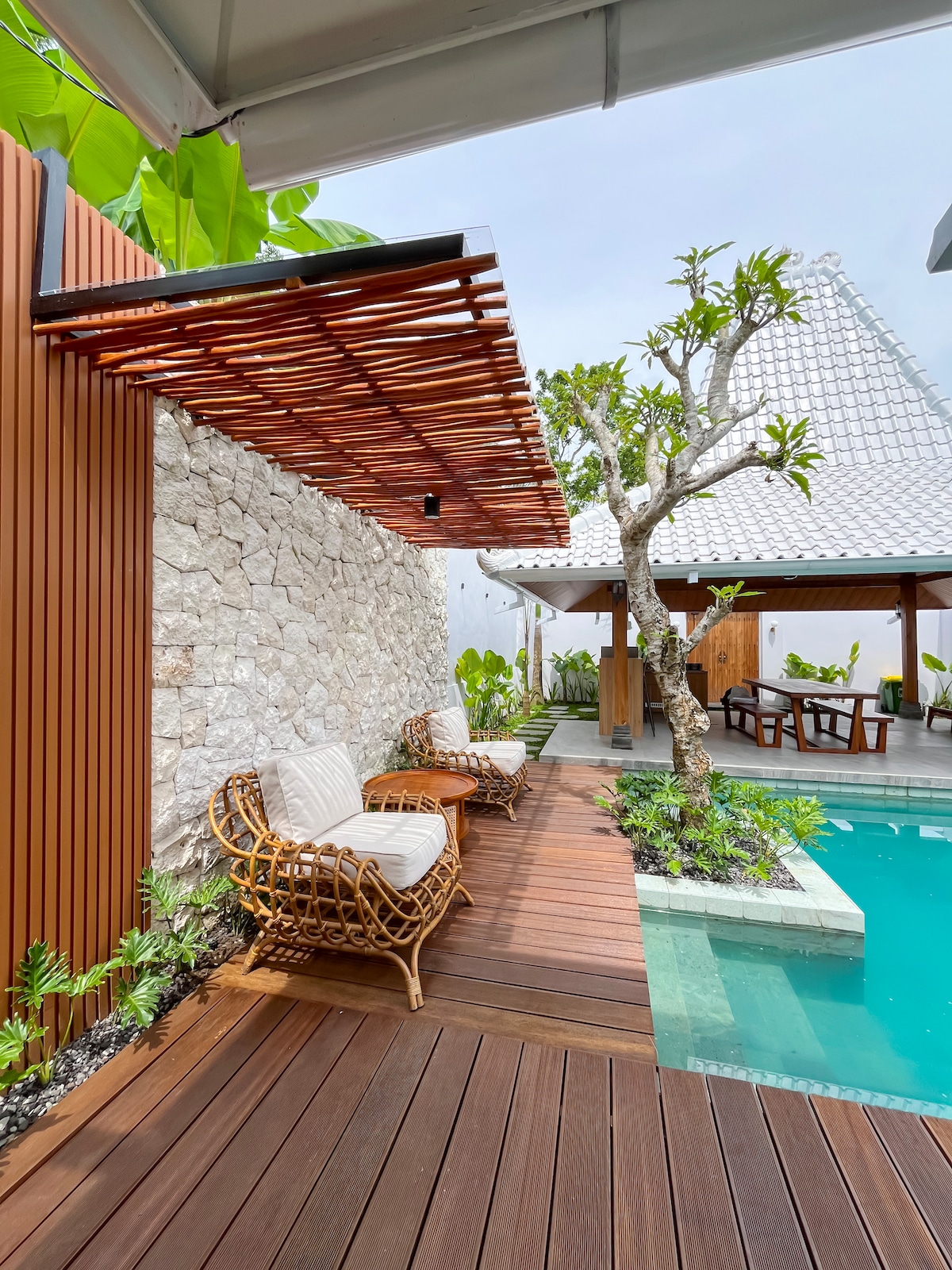 A serene outdoor space is presented with two wicker chairs positioned on a wooden deck, overlooking a private pool. Lush greenery and a small tree add natural elements, while a traditional Javanese roof is visible in the background, maintaining a connection to the villa's architectural heritage.