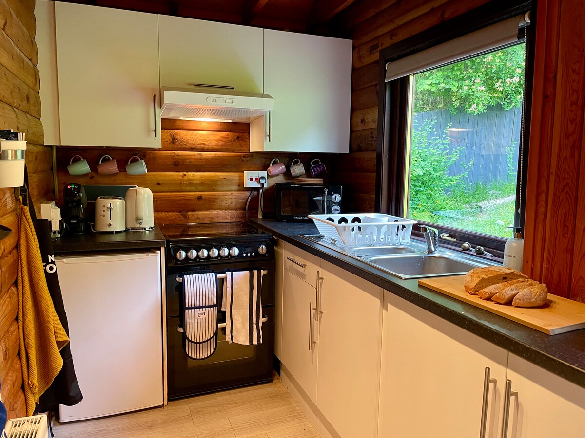 A modern kitchen is displayed, featuring light wood paneling and sleek cabinetry. Essential appliances, such as a refrigerator, stove, and a kettle, are visible. A large window brings natural light into the space, while a wooden cutting board is placed beside freshly sliced bread.