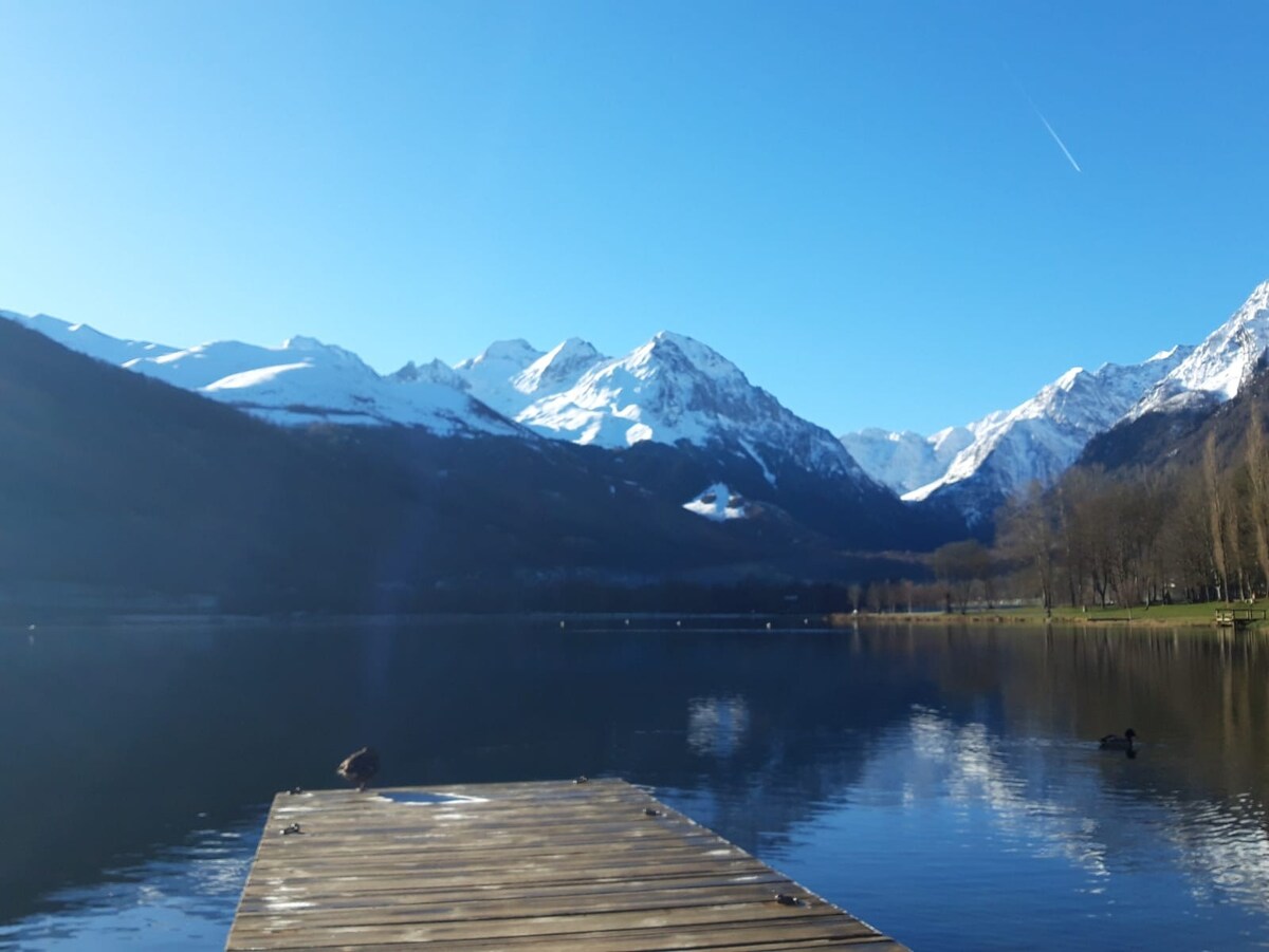 A wooden dock extends over a calm lake, reflecting the surrounding snow-capped mountains. The clear blue sky is visible above, while a few ducks can be seen on the water's surface. The serene landscape invites relaxation and outdoor exploration.