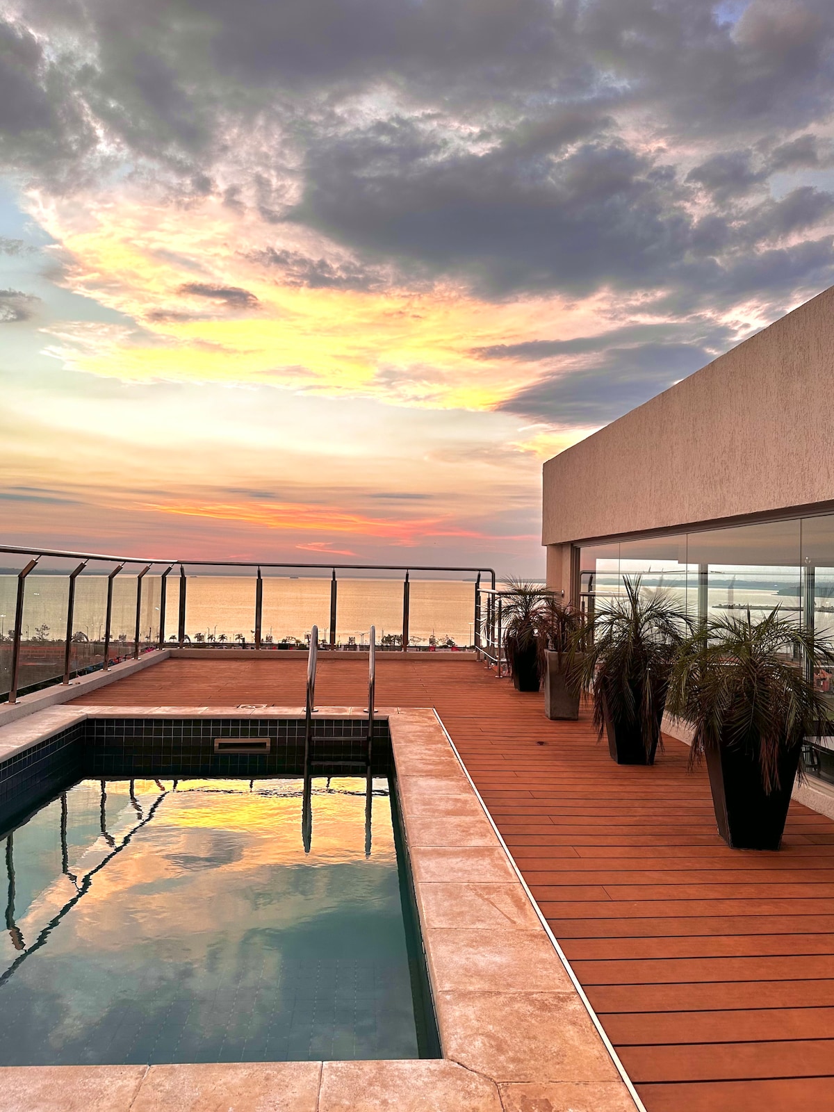 A rooftop terrace features a pool surrounded by wooden decking, with planters holding lush green plants. The horizon showcases a vibrant sunset over the sea, reflecting orange and pink hues in the water, while the modern building overlooks this serene scene.