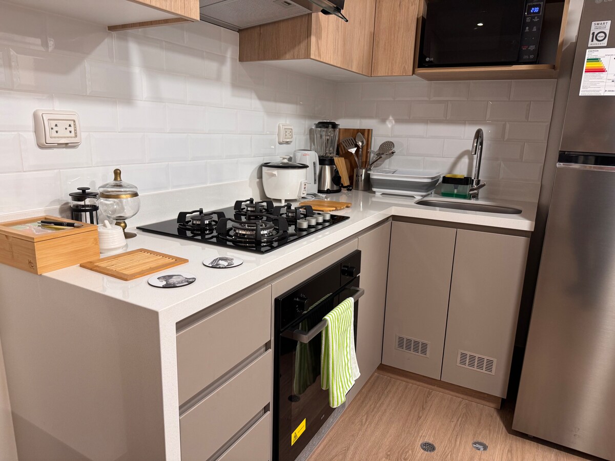 The kitchen area includes modern cabinetry and a sleek countertop. A gas stove is situated below a range hood, with a microwave above. Various kitchen appliances, including a coffee maker and kettle, are visible. A sink with a dish rack is positioned alongside storage spaces.