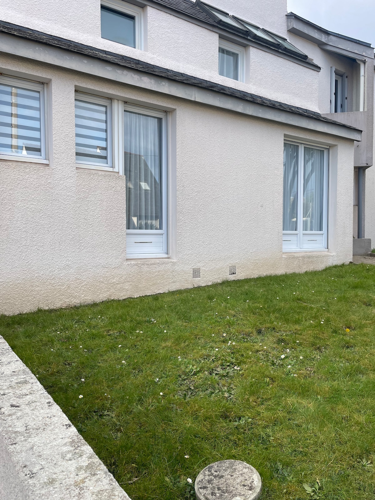 A grassy outdoor area is featured, bordered by a low stone wall. Two large windows are visible, providing light to the interior. The walls are finished in a light color, complementing the green lawn and the overall simple exterior design.