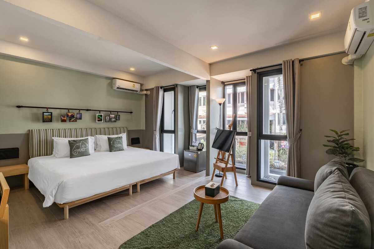 The room features a spacious bed with light bedding and a comfortable sofa next to a small coffee table. Large windows allow ample natural light, complemented by soft grey curtains. A small desk with an easel is positioned alongside a greenery accent for a touch of nature.