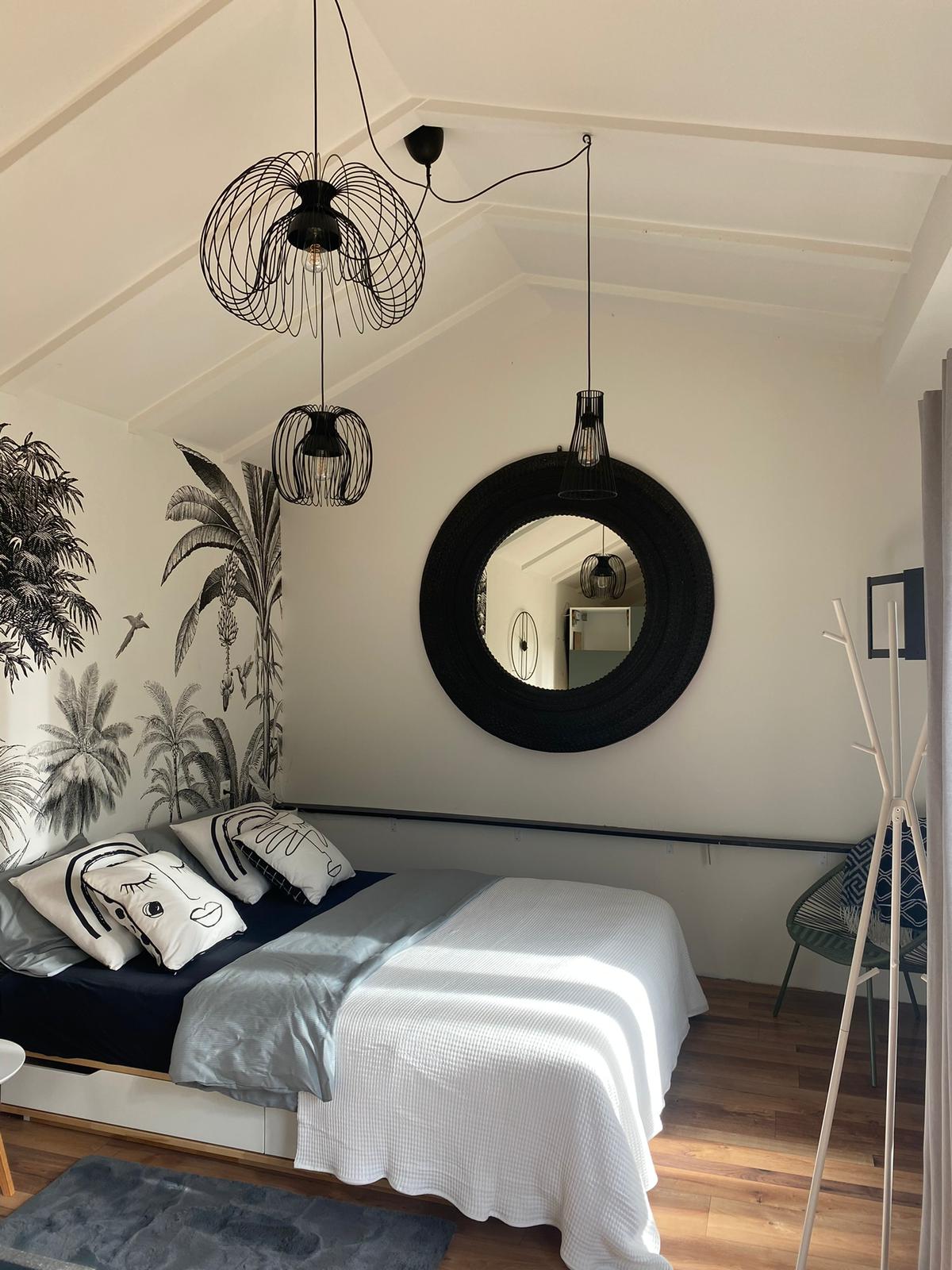 A sleeping area features a spacious bed with light bedding. Decorative pillows with abstract designs are arranged on the bed. Unique ceiling lights hang above, and a large mirror reflects part of the room. Tropical wallpaper adds an artistic touch to the space.