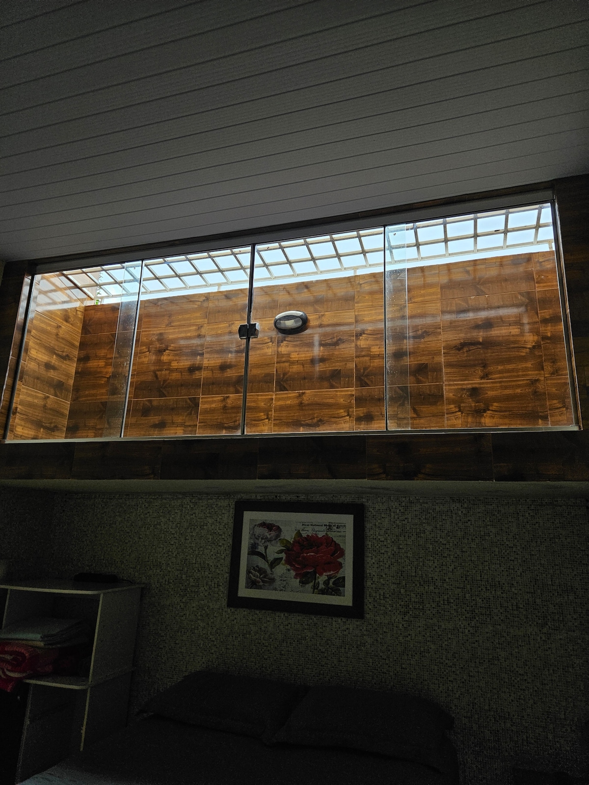 A large glass panel is positioned above the bed, allowing natural light to illuminate the room. The wooden wall behind the glass adds warmth to the space. A decorative floral piece is visible on the wall, enhancing the room's overall design.