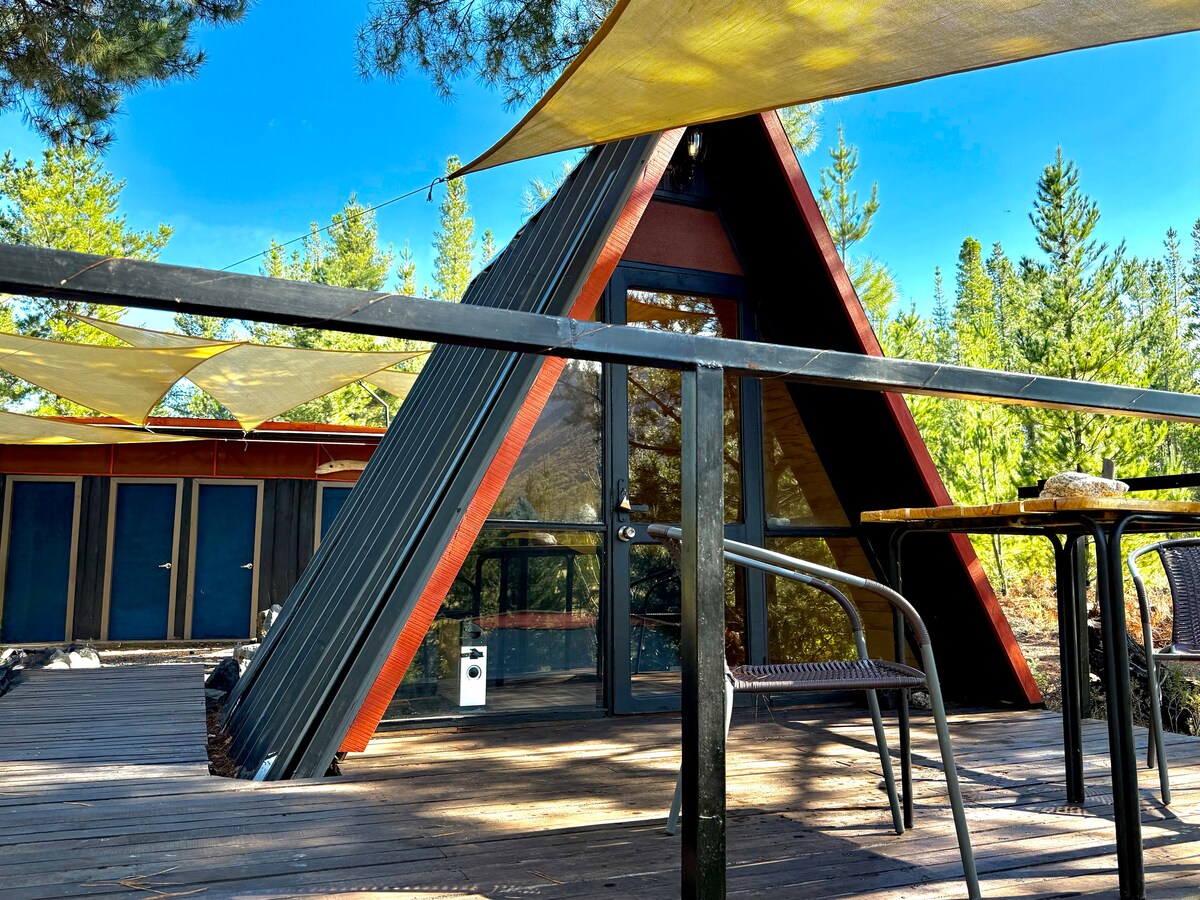 An A-frame cabin is seen from the deck, surrounded by trees. The structure features large glass windows, allowing natural light to flow inside. Sun shades are stretched above the deck, and outdoor seating is provided with a small table and chairs, inviting relaxation in nature.