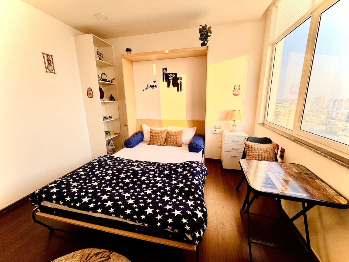 A cozy bedroom features a spacious bed adorned with a star-patterned blanket. Natural light floods the room through large windows, illuminating the nearby workspace and decorative shelves. A bedside lamp adds a touch of warmth, complementing the serene ambiance.