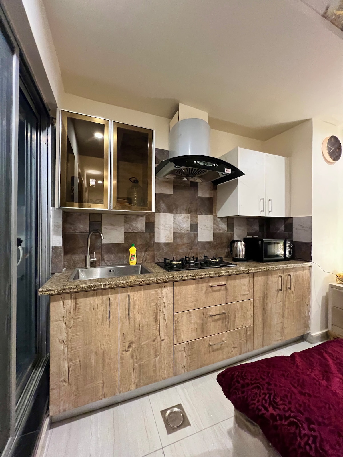 A compact kitchen area features wooden cabinetry and granite countertops. A stainless steel sink and gas stove are included, alongside a microwave and kettle. Neutral-colored tiles decorate the backsplash, and a window allows natural light into the space.