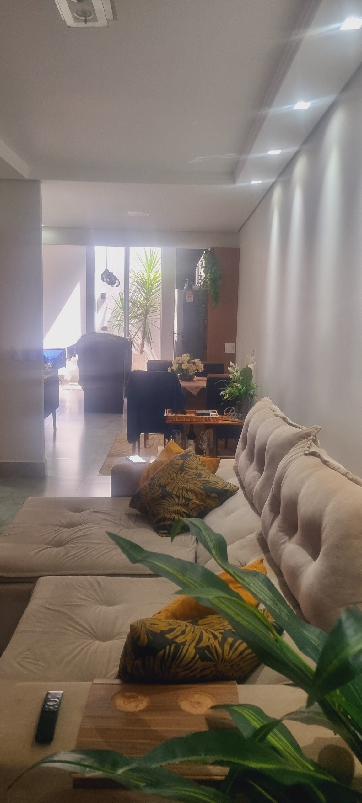 A cozy living area features a beige sectional sofa adorned with decorative cushions. A coffee table is positioned in front, complemented by a small potted plant. In the background, natural light filters through large glass doors, illuminating the adjoining dining space.