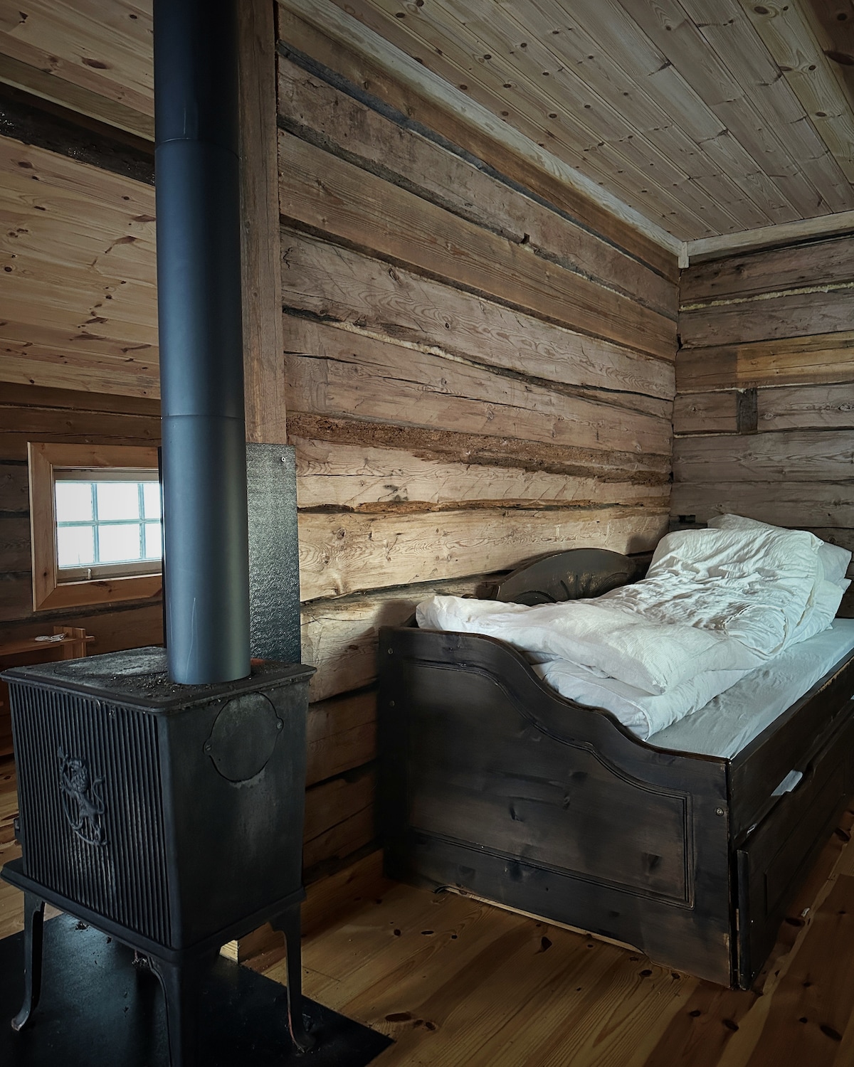 A rustic sleeping area features a wooden bed with a simple white bedding arrangement. The room is surrounded by the natural texture of light wood walls, and a cast-iron stove stands nearby, providing warmth.