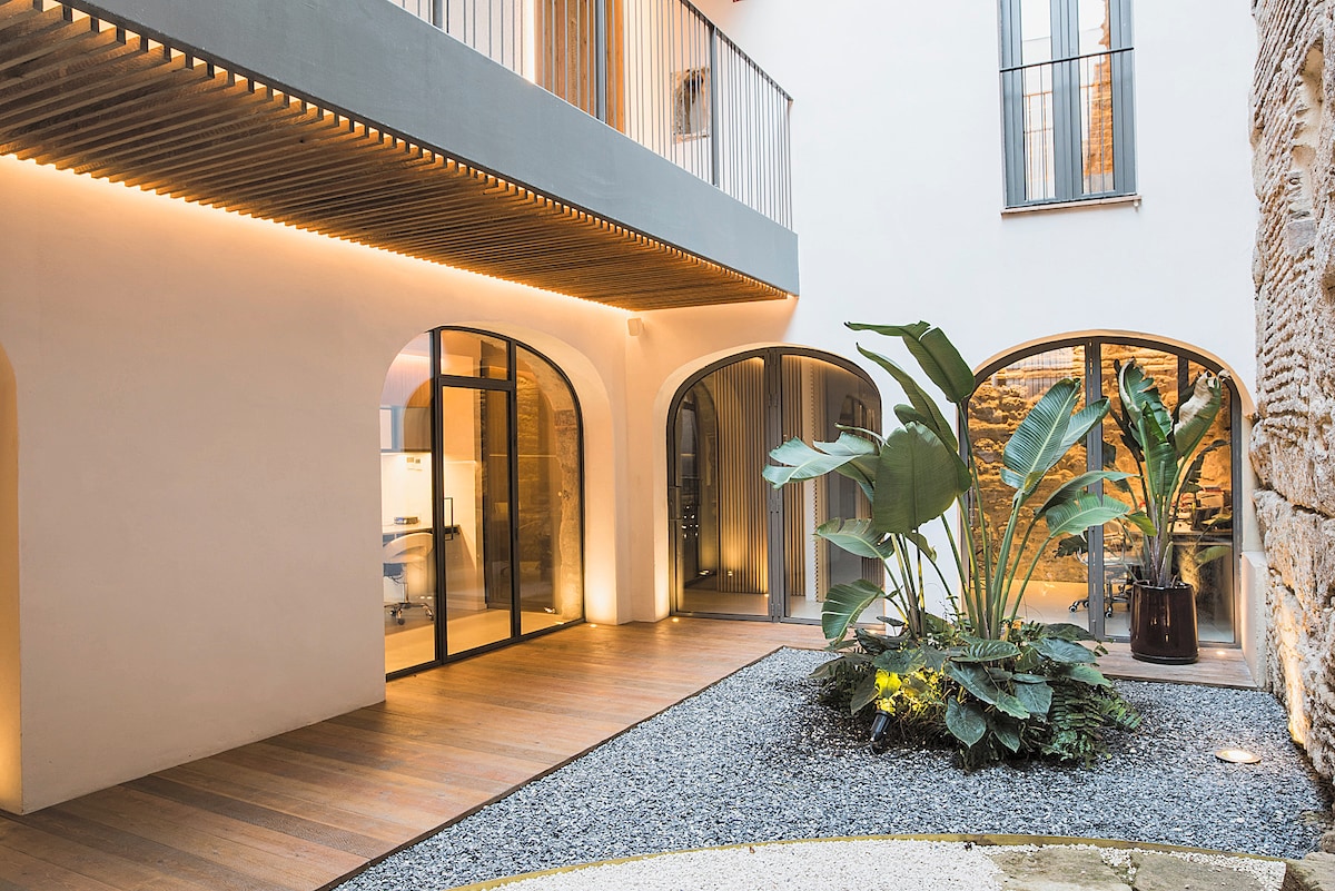 An inviting courtyard showcases smooth wooden flooring bordered by sleek, curved arches. Lush greenery is present, with large potted plants positioned around a central landscaped area of gravel and foliage. Soft lighting enhances the serene ambiance, inviting relaxation and exploration.