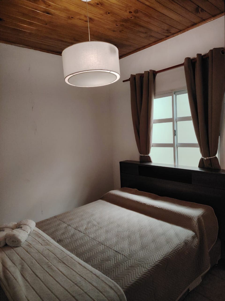 A cozy bedroom features a spacious bed with a textured blanket and a rolled towel nearby. Natural light filters through a window, while soft curtains frame the view. A modern ceiling light illuminates the room, and wooden paneling adorns the ceiling.
