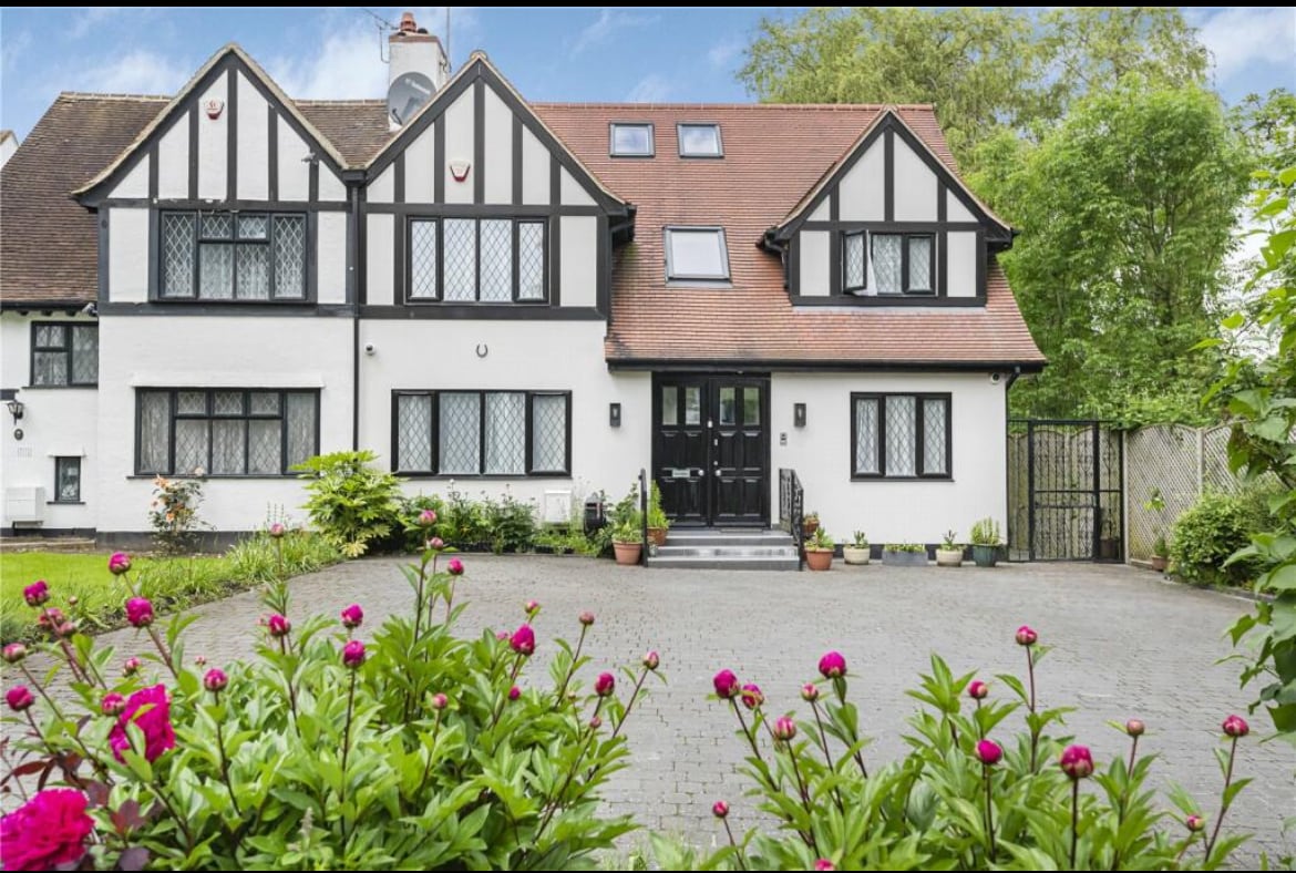 A two-story Tudor-style house exhibits a combination of white and black timber framing. The entrance is framed by a small landscaped area with flowering plants. An inviting pathway leads to the main door, with windows adorned in a classic diamond pattern, enhancing the building's architectural appeal.
