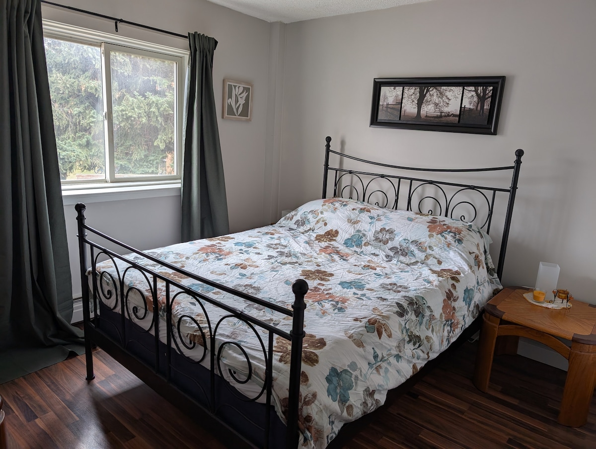 A comfortable sleeping area is presented, featuring a spacious bed with a floral-patterned bedding set. A nightstand is positioned beside the bed, holding a few items. Natural light fills the room through a large window, framed by dark curtains that complement the decor.