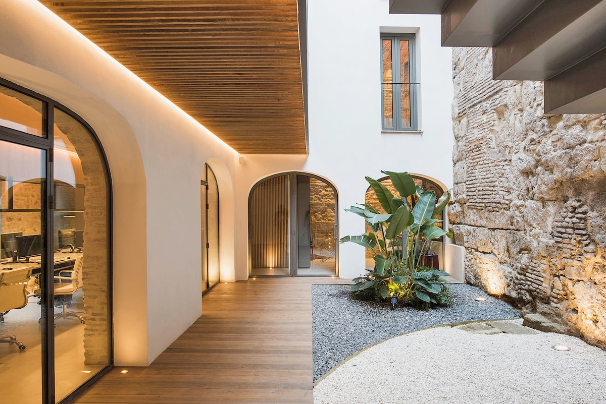 An open courtyard features a blend of modern and historic elements. Smooth wooden decking transitions to gravel pathways surrounded by lush plants. Natural light filters through large glass doors, enhancing the unique architectural details of the stone walls and creating a serene outdoor space.