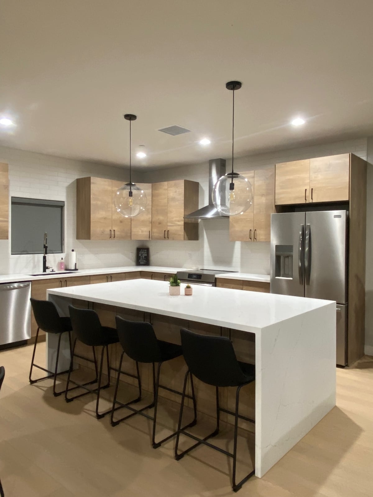 A modern kitchen showcases wood-grain cabinetry paired with sleek stainless steel appliances. A spacious island with a white countertop provides seating for four. Pendant lights hang above, illuminating the preparation area while a large window allows for natural light.
