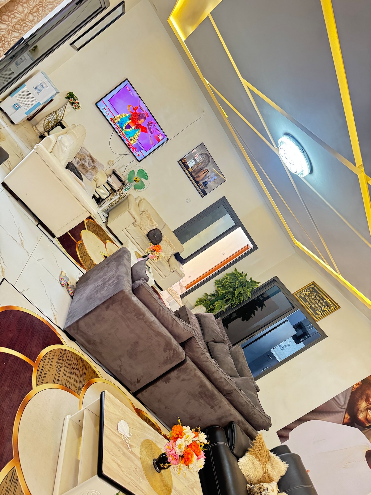 A spacious living area features a large, comfortable sectional sofa seating area, facing a wall-mounted television. A coffee table with decorative elements sits in the center, surrounded by light beige upholstered furniture. Soft lighting highlights the unique ceiling design, enhancing the room's layout.