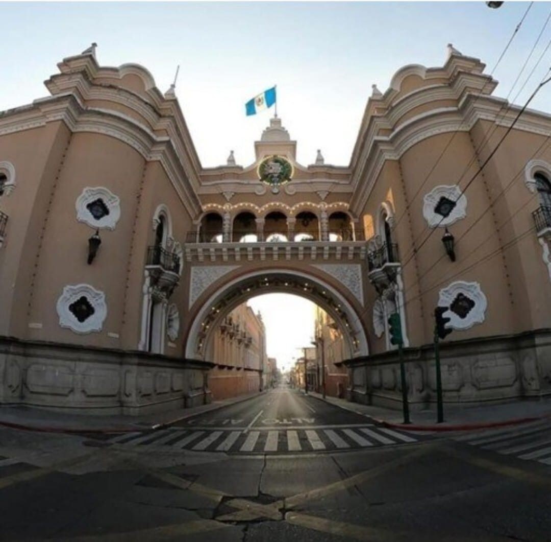 An iconic archway of a historical building is prominently featured, with the Guatemalan flag displayed at the top. The surrounding street is empty, showcasing a wide crosswalk and distinct architectural details of the building's facade, which includes decorative elements and balconies.