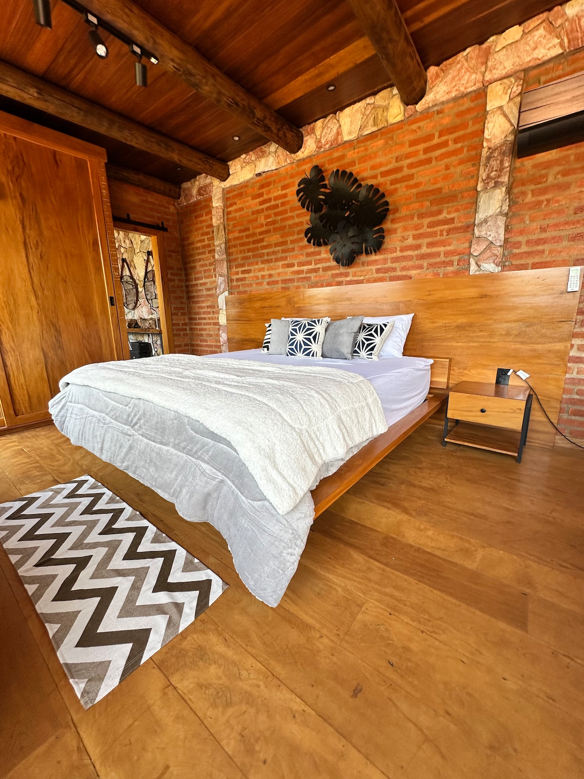 A wooden bed is centered in the room, featuring a simple gray blanket laid over fresh white linens. Decorative cushions with geometric designs are placed against the headboard. A bedside table holds a lamp, and a patterned rug adds texture to the wooden floor.