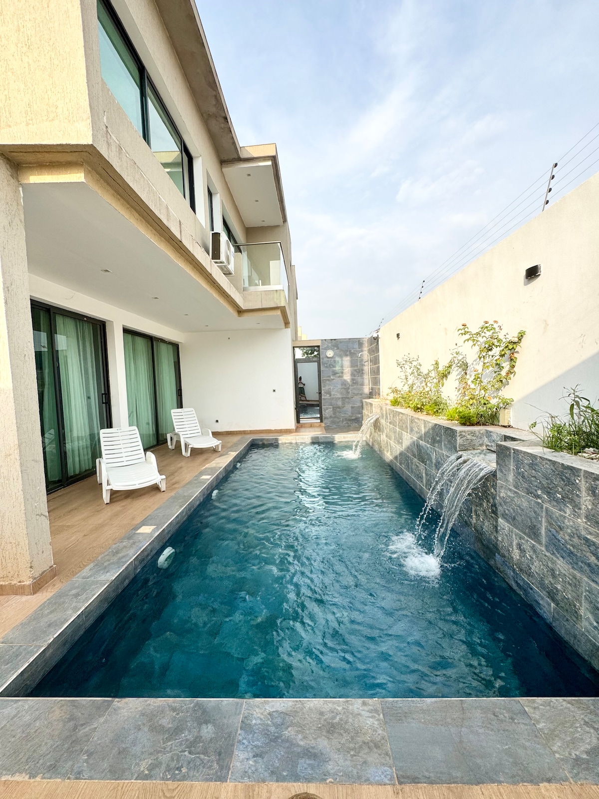 A serene swimming pool is featured alongside the house, framed by sleek tiles and a small water feature. Two lounge chairs are arranged nearby, providing space for relaxation. The space is bathed in natural light, with large glass doors offering views of the pool area.