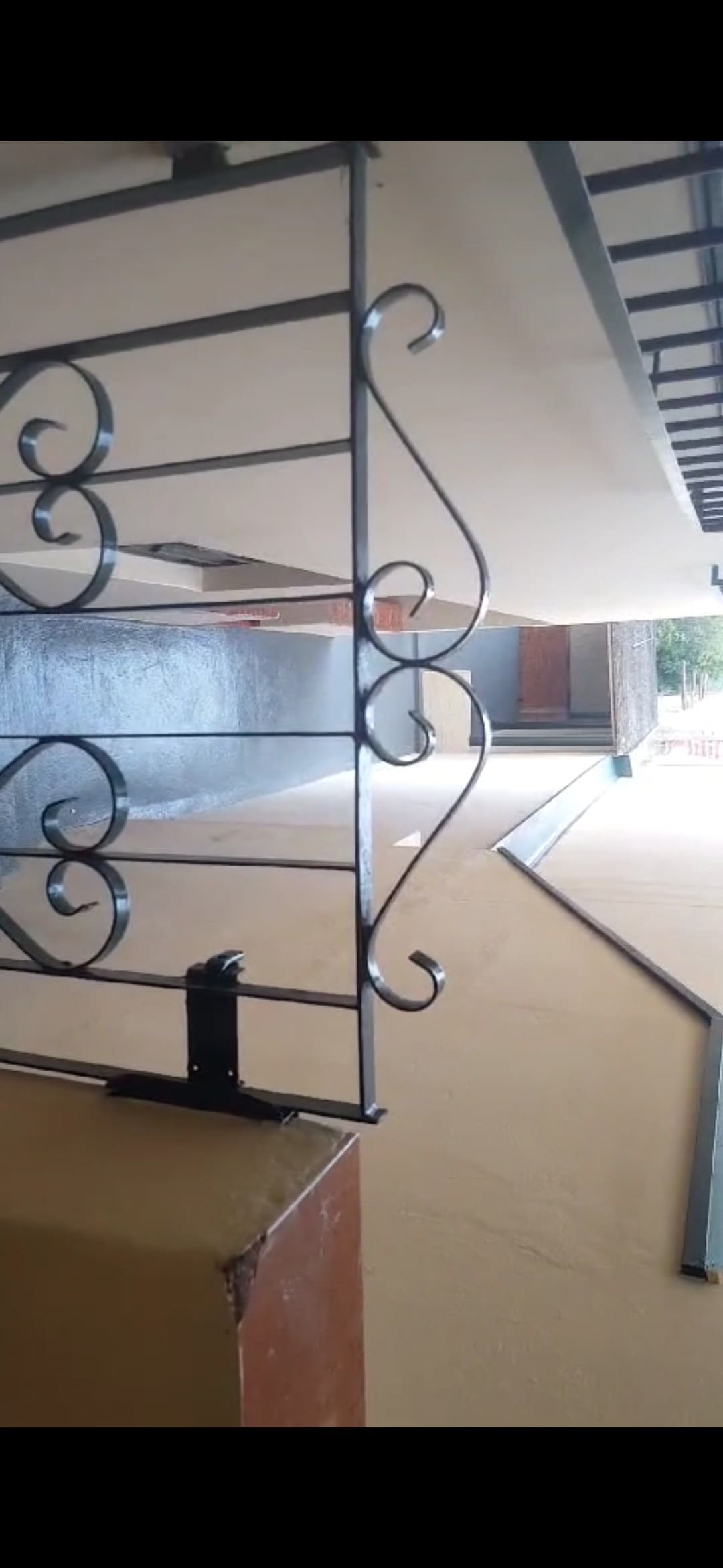 A metal balcony railing showcases an intricate design, creating an open view into a covered walkway. The muted color of the walls and ceiling complements the simplicity of the space, with natural light filtering in from the upper area.