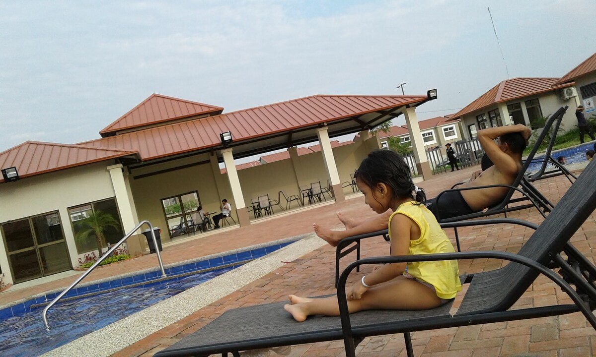 A serene poolside scene showcases children relaxing on lounge chairs. The area features a tranquil pool with a textured stone edge. Nearby, covered seating is visible, offering shaded options. Charming buildings with terracotta roofs create a welcoming backdrop against a softly clouded sky.
