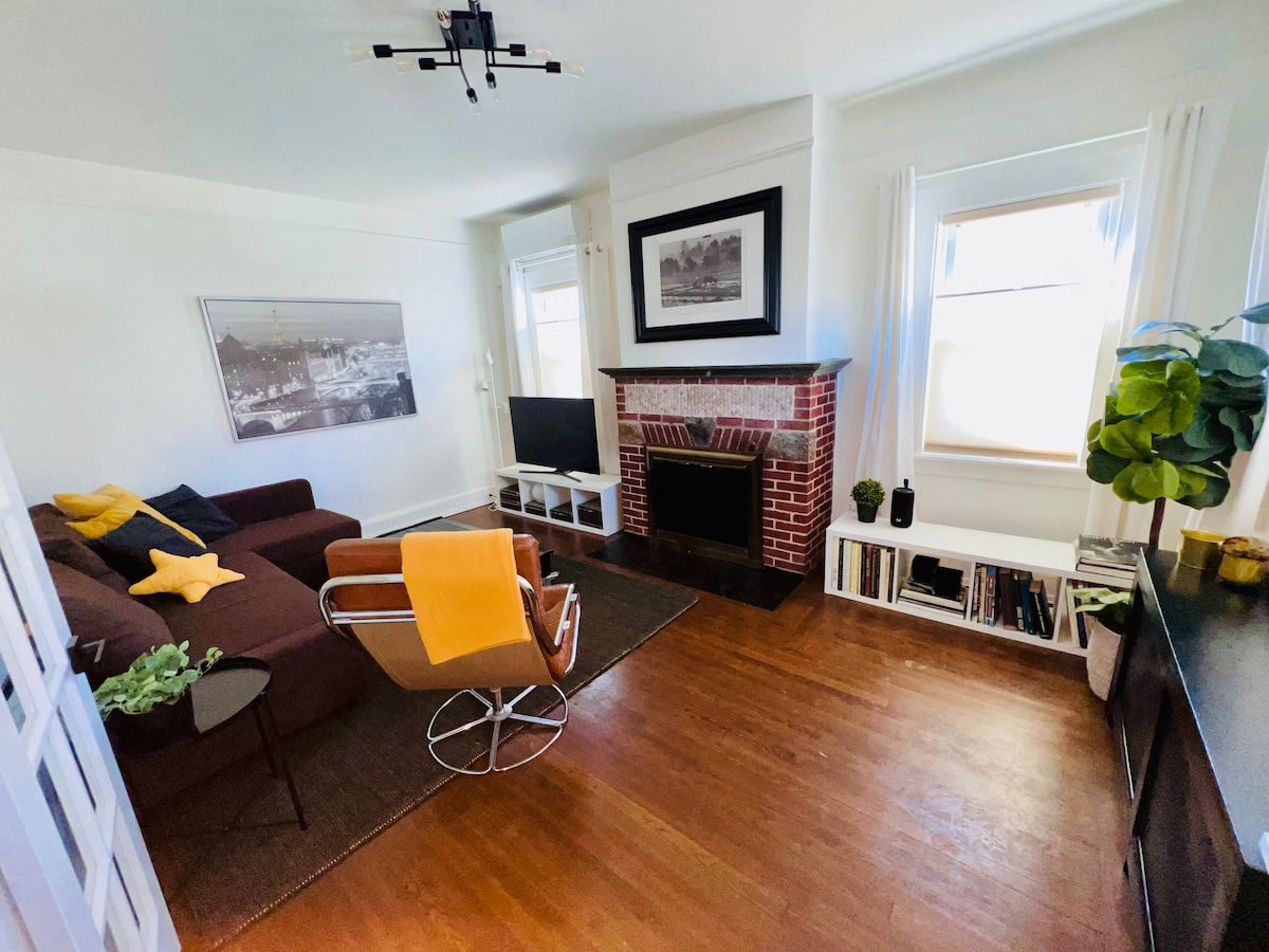 A living room features a brown sofa with yellow and gray accent pillows, paired with a sleek armchair. A flat-screen TV is mounted on a white media unit, with bookshelves and decorative plants enhancing the space. A brick fireplace adds a classic touch to the room.